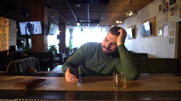 Un hombre barbudo está sentado en un bar bebiendo whisky y enviando mensajes de texto por teléfono con una novia — Vídeos de Stock