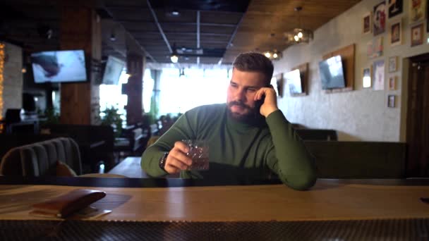En skäggig man sitter i en bar och dricker whisky och pratar i telefon. — Stockvideo
