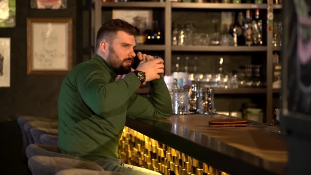 Een man met een baard drinkt alcohol en praat aan de telefoon. — Stockvideo