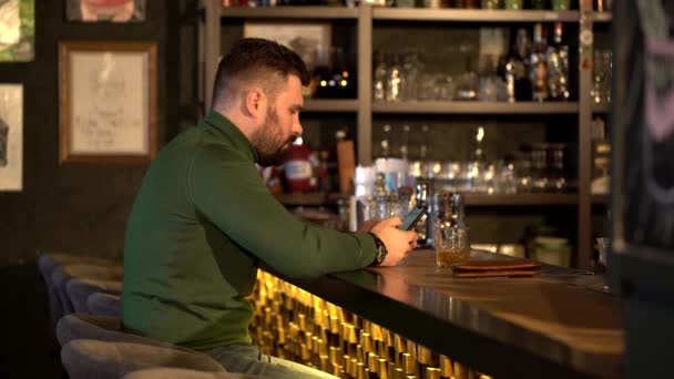 El tipo se sienta en el mostrador del bar con vasos de alcohol . — Vídeos de Stock