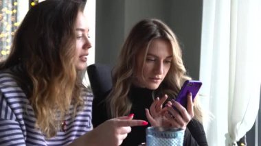 İki bayan arkadaş kafede oturuyor ve cep telefonundan fotoğraf izliyorlar.