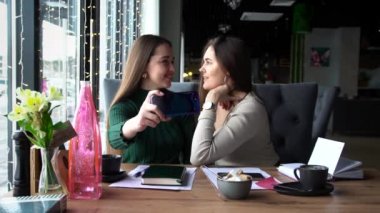Mutlu iki kadın gülümser ve kameranın önünde poz verir, fotoğraf çekmek için modern cep telefonunu kullanır.