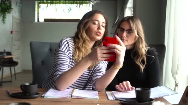 Jovens meninas alegres melhores amigos estão sentados em um café, usando um smartphone e rindo — Vídeo de Stock