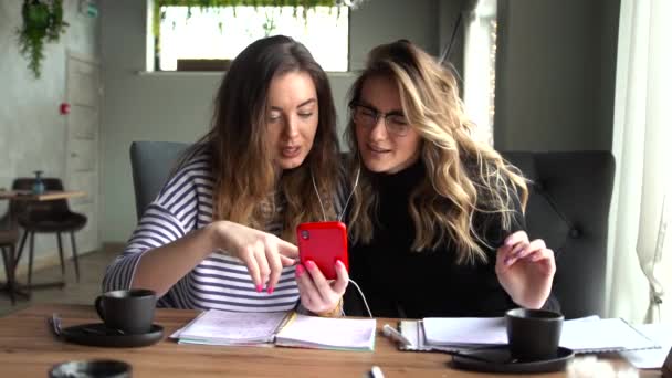 Two women sit in a cafe during the day and watch a video together on a smartphone. — Stock Video