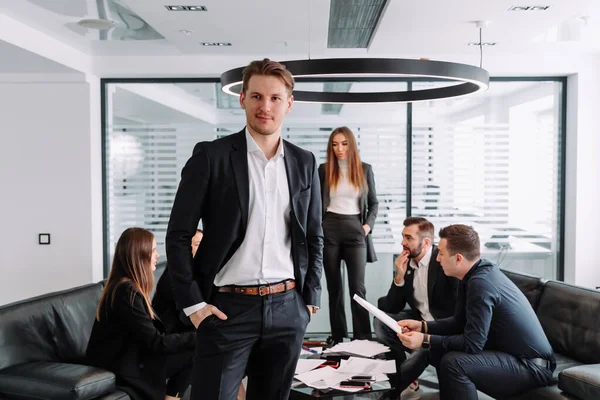 Mutlu iş adamının portresi, kameraya bakıp gülümsüyor. İş ekibi arka planda çalışıyor.