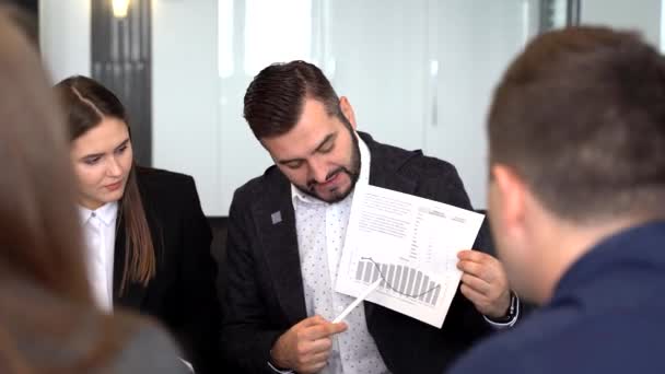 Ein gutaussehender junger Mann zeigt auf eine Grafik, während seine Kollegen zuhören und an einem Tisch sitzen — Stockvideo