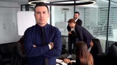 Arkaplanda iş arkadaşlarıyla konuşurken kollarını kavuşturmuş kendinden emin genç bir adam.