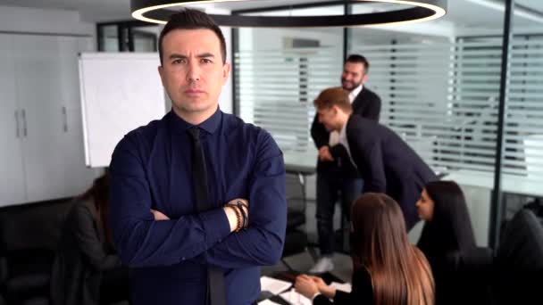 Confident young man holding arms folded while standing at the table with talking colleagues on a background — Stock Video