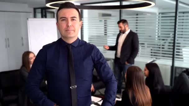 Důvěryhodný mladý muž drží ruce složené, zatímco stojí u stolu s mluvícími kolegy na pozadí — Stock video