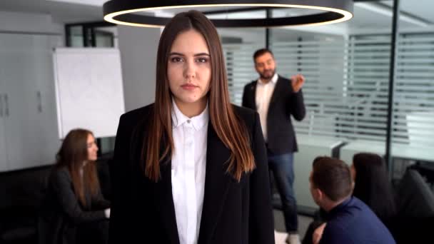 Деловая женщина в современном открытом плане офисное рабочее пространство улыбается в камеру с коллегами на заднем плане — стоковое видео