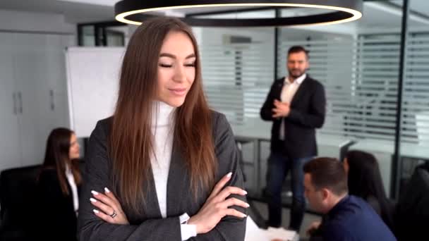 Fiatal bájos üzletasszony néz a kamera előtt az irodában, mosolyog, kollégái hálózatba szerveződnek a technológiákkal — Stock videók
