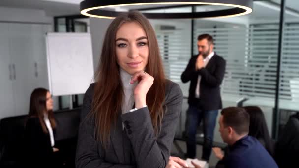 Egy fiatal üzletasszony szürke öltönyben pózol a kamerának, kollégák homályos háttérrel dolgoznak — Stock videók