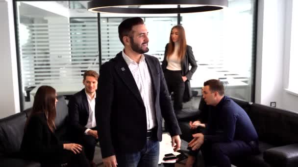 Zelfverzekerde jongeman houdt armen gevouwen terwijl hij aan tafel staat met pratende collega 's op een achtergrond — Stockvideo