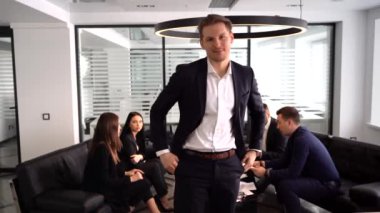 Arkaplanda iş arkadaşlarıyla konuşurken kollarını kavuşturmuş kendinden emin genç bir adam.