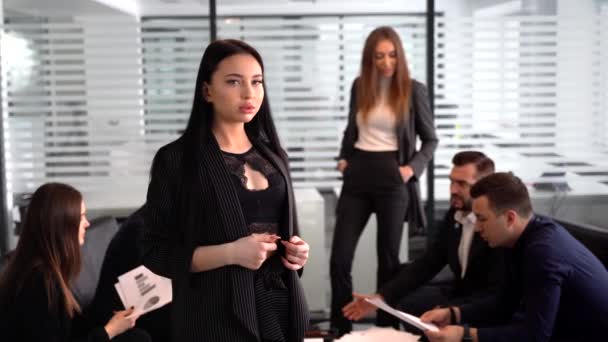 Glimlachende zakenvrouw in het kantoor, handen gevouwen en zelfverzekerde uitdrukking als andere werknemers houden een vergadering op de achtergrond — Stockvideo