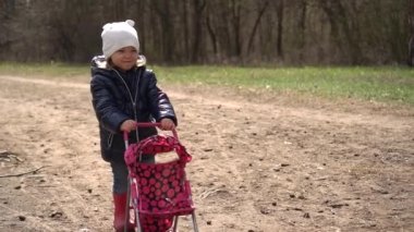 Mutlu çocuk çimenlerde oynuyor. Güzel küçük kız dışarıda bir bebek arabasıyla oynuyor. Sür bebek arabasını, mutlu kız bebek. Yavaş çekimde.