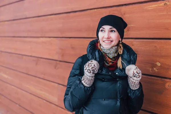 Young smiling woman in winter cloths standing in front of wooden wall outdoors