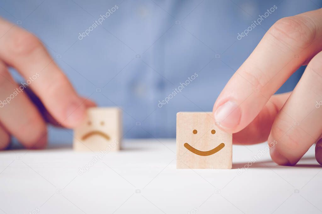 A man holds with his fingers a wooden cube with a positive face next to a disgruntled one. Concept for evaluating an action or resource.