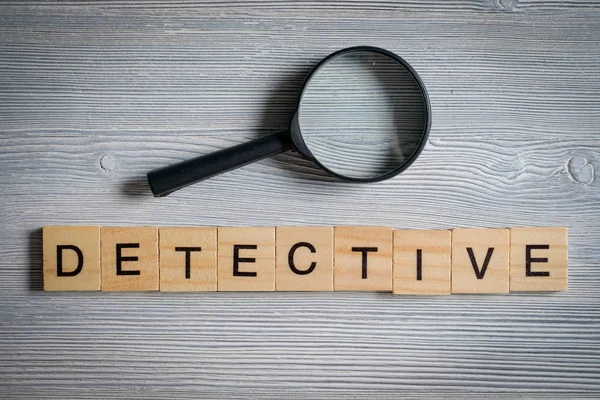 Ord av trä bokstäver - detektiv, på en grå bakgrund med en förstoringsglas. Begreppet undersökande yrke. — Stockfoto