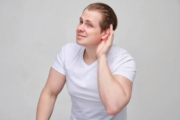 Un joven con una camiseta blanca no puede oír bien. La mano está cerca de la oreja . —  Fotos de Stock