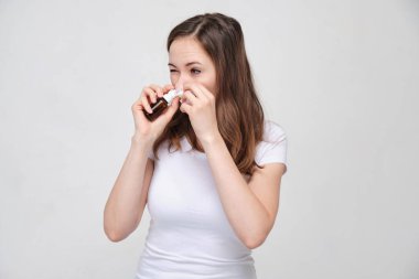 Beyaz tişörtlü, gri arka planda çekici bir kız. Burnunda tıbbı eziyor. Soğuk algınlığı ve gribin önlenmesi ve tedavisi.