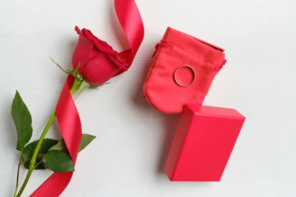 Una hermosa rosa roja con una cinta al lado de la caja y un anillo de oro sobre un fondo de madera blanca. Vista superior . — Foto de Stock
