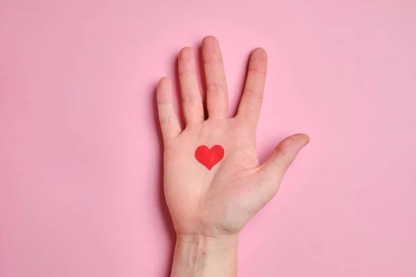 In de palm van de hand zit een klein rood hartje op een achtergrond van roze papier. — Stockfoto