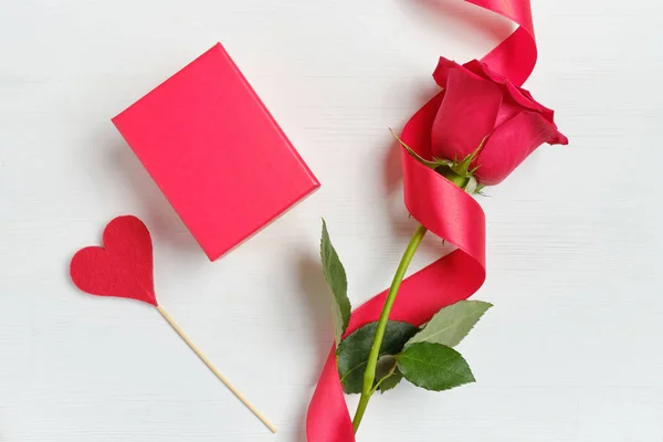Una hermosa rosa roja con una cinta al lado de la caja sobre un fondo de madera blanca. Vista superior . —  Fotos de Stock