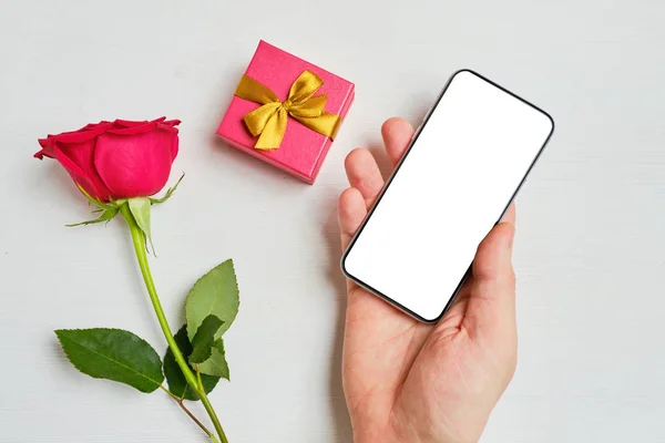 A kéz tartja az okostelefon fénymásolás mellett az ajándék doboz és a vörös rózsa egy fehér fa háttér. — Stock Fotó