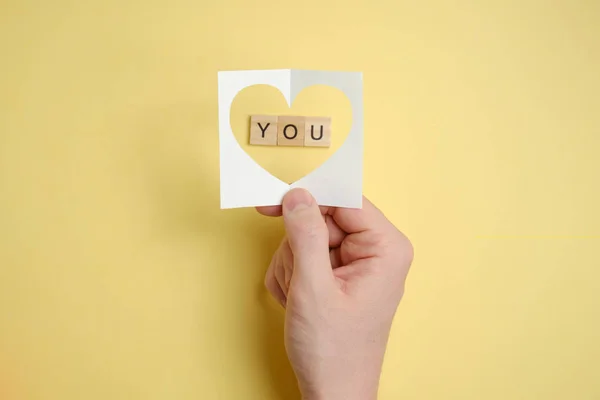 Een liefdesverklaring. Hand houdt abstracte papieren hart over houten vierkanten met het woord - u. Papieren gele achtergrond. — Stockfoto