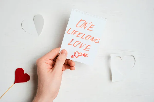 Een hand houdt een stuk papier met een rode inscriptie - in liefde - naast abstracte harten op een houten witte achtergrond. — Stockfoto