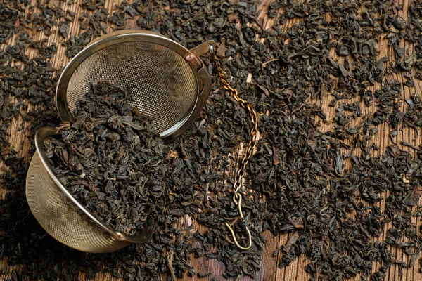 Black tea in strainer with chain on scattered tea on wooden background. — 图库照片