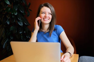 Happy beautiful girl sits in a cafe and talks on the phone in front of a laptop.