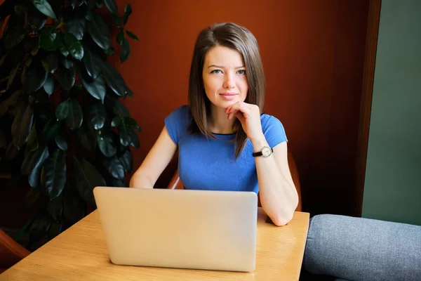 Ένα όμορφο κορίτσι κάθεται σε ένα καφέ ντυμένο με ένα μπλε φόρεμα πίσω από ένα φορητό υπολογιστή. Έννοια freelancer — Φωτογραφία Αρχείου