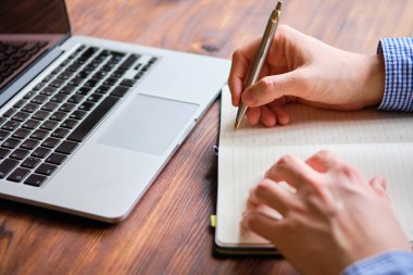 A man makes notes in a diary next to a laptop on a wooden table.