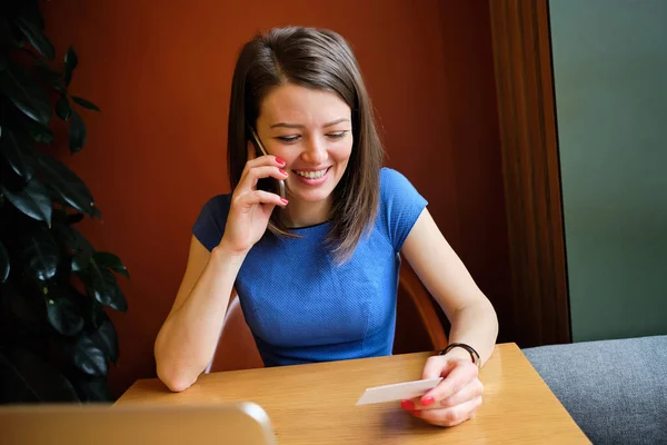 Χαμογελώντας κορίτσι σε ένα καφέ καλώντας σε ένα smartphone, ενώ κοιτάζοντας μια επαγγελματική κάρτα με επαφές. — Φωτογραφία Αρχείου