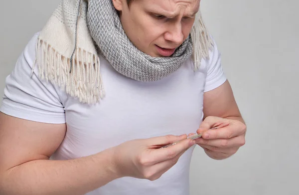 A man in a white T-shirt with a scarf looks at the thermometer in surprise. The concept of lowered body temperature. — Stockfoto