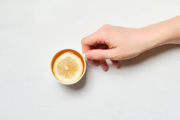 Tangan itu memegang secangkir teh dengan lemon di latar belakang kayu putih. Tampilan atas . — Stok Foto