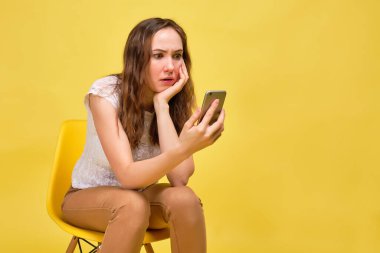 A girl in a white T-shirt and brown jeans on a yellow background is in surprise looking at new email messages. Spam concept.