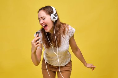 A girl in a white T-shirt and brown jeans on a yellow background depicts that she sings along to the music in a smartphone like a microphone and dances.