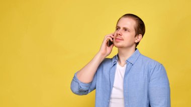 A man in a blue shirt on a yellow background is calling on his smartphone.