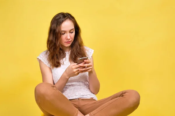 Girl in a white T-shirt and brown jeans on a yellow background prints a message in a smartphone. — 图库照片