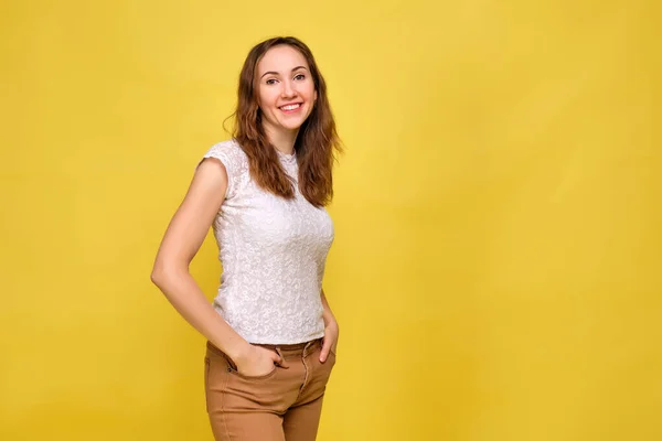 Een meisje in een wit T-shirt en bruine jeans op een gele achtergrond glimlacht en kijkt vrolijk naar de camera. — Stockfoto