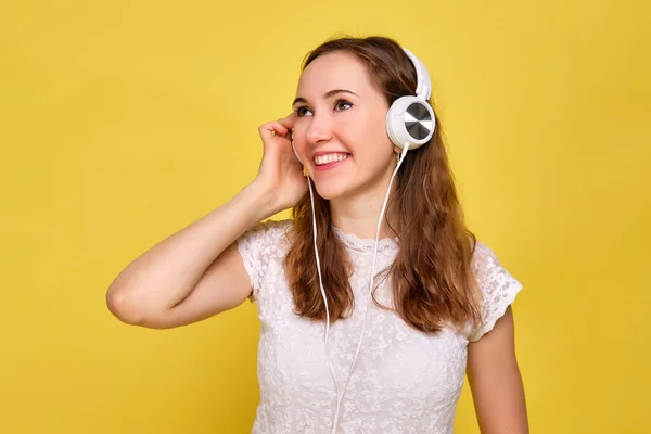 Una chica con una camiseta blanca y jeans marrones sobre un fondo amarillo relaja y escucha música con auriculares blancos . —  Fotos de Stock