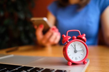 A girl dressed in a blue dress works in a smartphone in the foreground a red clock. Concept dead line.