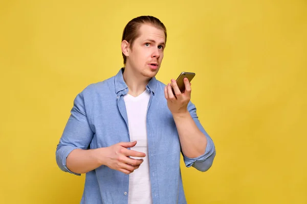 Ein Mann im blauen Hemd auf gelbem Hintergrund macht eine Audiobotschaft in einem Smartphone. — Stockfoto