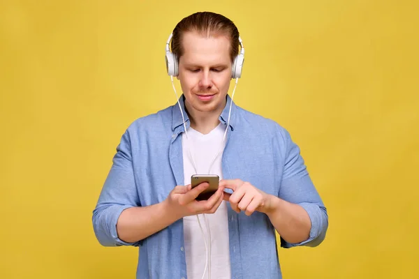 Muž v modré košili na žlutém pozadí v bílých sluchátkách poslouchá hudbu a přepíná písně v chytrém telefonu. — Stock fotografie