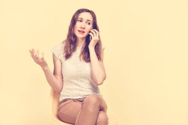 A brunette girl in summer clothes sits on a yellow background and speaks on the phone. Toned.