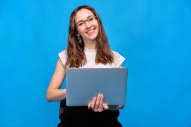 A brunette girl in casual clothes is holding a laptop on a blue background with glasses holding a laptop and looking at the camera.