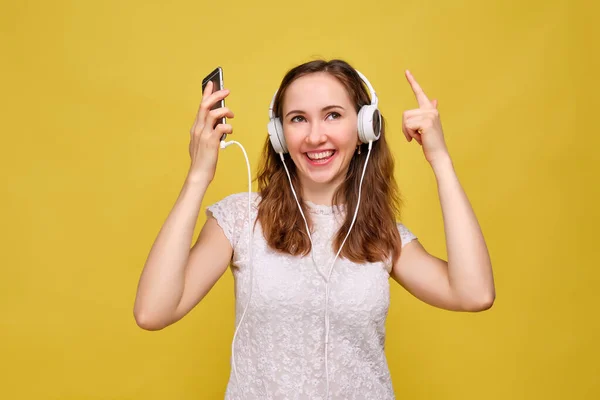 A brunette girl in summer clothes listens to music on headphones on a yellow background and dances. — Stock fotografie